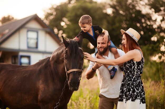 Glückliche junge Familie mit einem kleinen Sohn steht mit einem Pferd vor einem kleinen Landhaus