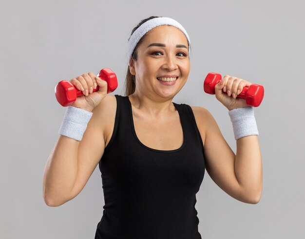 Glückliche junge Fitness-Frau mit Stirnband, die Hantel hält und Übungen macht, die selbstbewusst lächelnd über weißer Wand stehen?