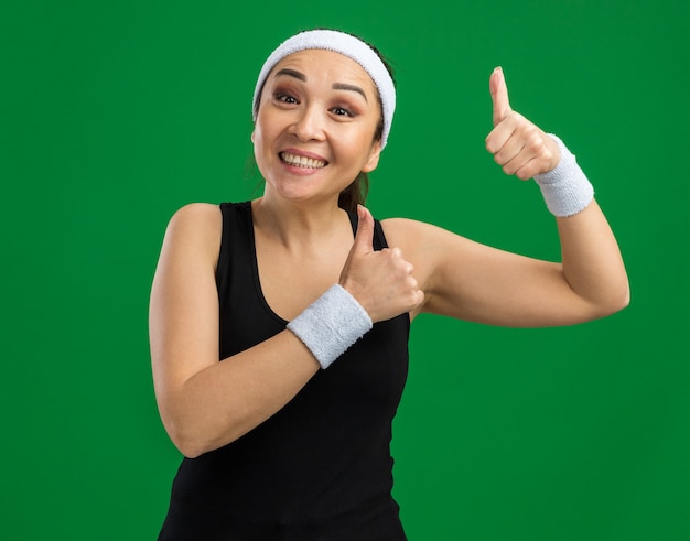Glückliche junge Fitnessfrau mit Stirnband und Armbinden mit einem Lächeln im Gesicht, das Daumen nach oben über grüner Wand zeigt