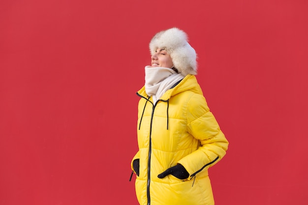 Glückliche junge Frau auf dem Hintergrund einer roten Wand in warmen Kleidern an einem sonnigen Wintertag