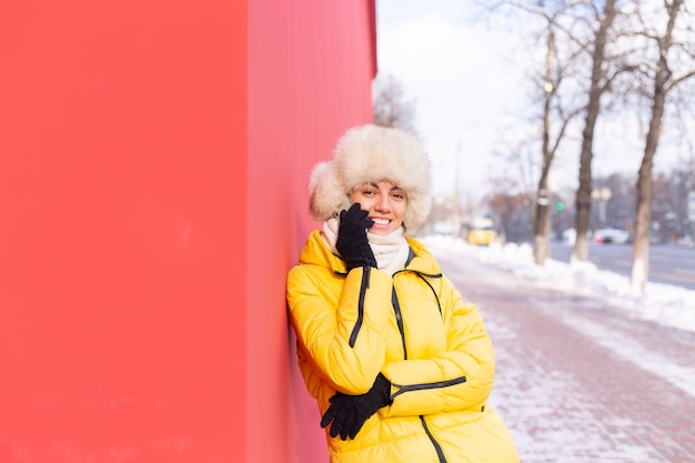 Glückliche junge Frau auf einem Hintergrund einer roten Wand in warmen Kleidern an einem sonnigen Wintertag lächelnd und am Telefon auf einem schneebedeckten Stadtbürgersteig sprechend