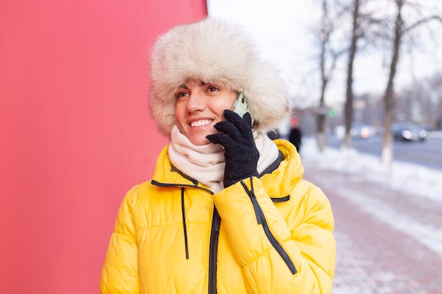 Glückliche junge Frau auf einem Hintergrund einer roten Wand in warmen Kleidern an einem sonnigen Wintertag lächelnd und am Telefon auf einem schneebedeckten Stadtbürgersteig sprechend