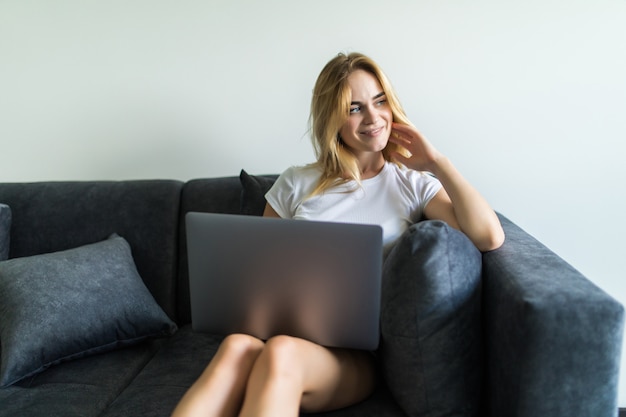 Glückliche junge Frau, die auf Couch mit ihrem Laptop zu Hause im Wohnzimmer sitzt