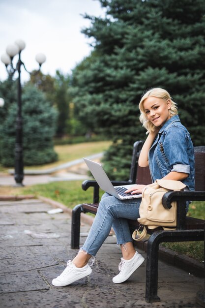 Glückliche junge Frau, die auf der Bank sitzt und Telefon und Laptop im Herbstmorgen der Stadt benutzt