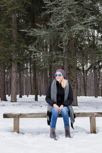 Glückliche junge Frau, die auf der Holzbank trägt warme Kleidung im Winter sitzt