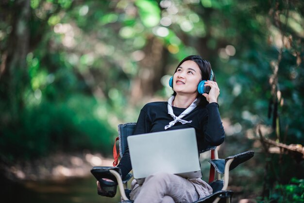 Glückliche junge Frau, die auf einem Campingstuhl sitzt und Kopfhörer trägt und Musik vom Laptop hört, während sie sich beim Camping im Wald entspannt