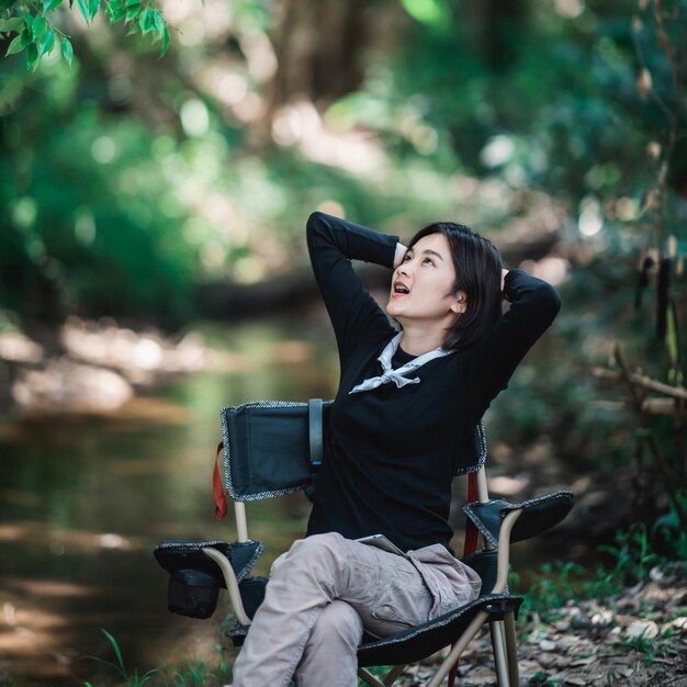 Glückliche junge Frau, die auf einem Stuhl sitzt, während sie sich beim Camping im Waldkopierraum entspannt