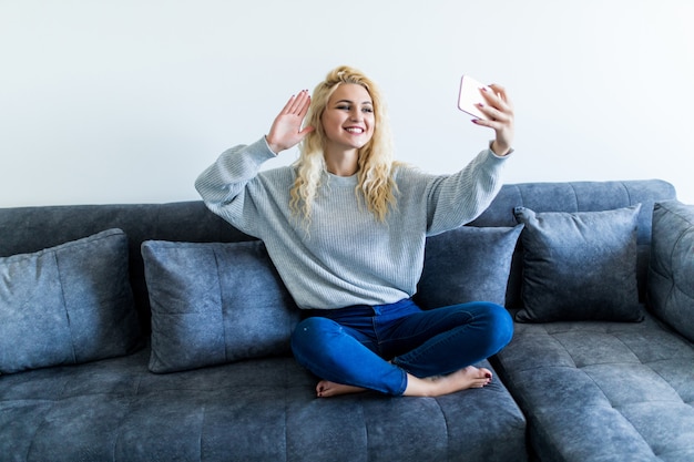 Glückliche junge Frau, die auf Sofa mit Telefon sitzt und Videoanruf zu Hause hat