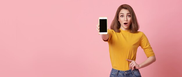 Glückliche junge Frau, die bei leerem Bildschirm-Handy und Handgestenerfolg einzeln auf rosa Hintergrund zeigt.