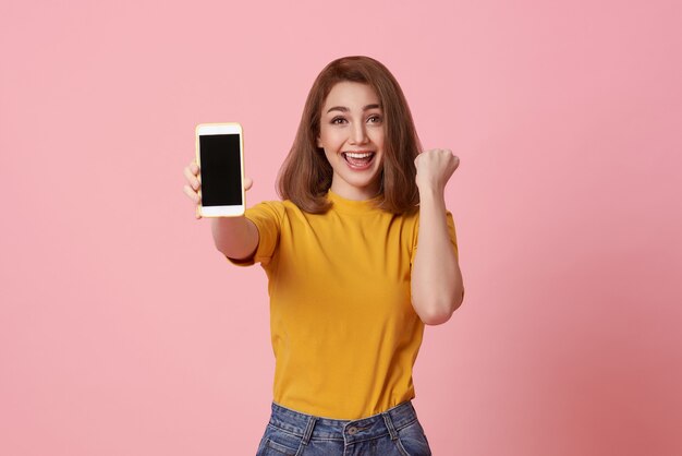Glückliche junge Frau, die bei leerem Bildschirm-Handy und Handgestenerfolg einzeln auf rosa Hintergrund zeigt.