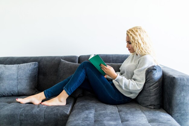 Glückliche junge Frau, die Buch auf Couch zu Hause liest
