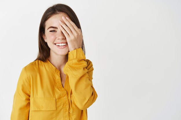 Glückliche junge Frau, die den Anblick überprüft, die Hälfte des Gesichts mit der Hand bedeckt und lächelt, mit einem Auge nach vorne schaut und über der weißen Wand steht.