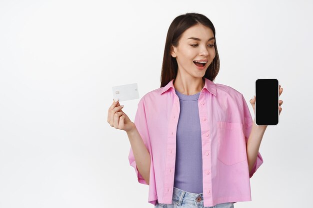 Glückliche junge Frau, die den Smartphone-Bildschirm mit der Kreditkarte sieht und zeigt Konzept der mobilen Zahlung beim Online-Shopping oder Kauf auf weißem Hintergrund