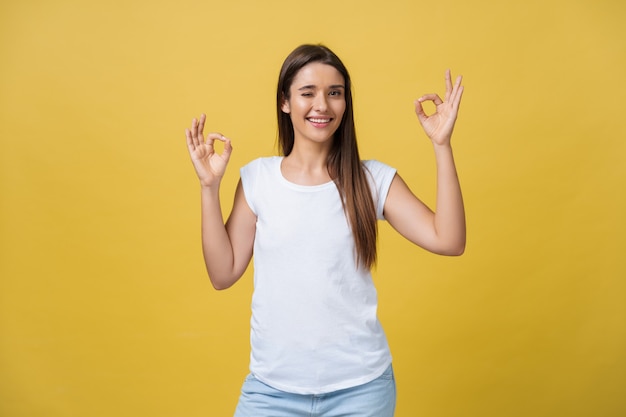 Glückliche junge Frau, die ein Okay-Zeichen mit den Fingern zeigt und auf einem gelben Hintergrund zwinkert