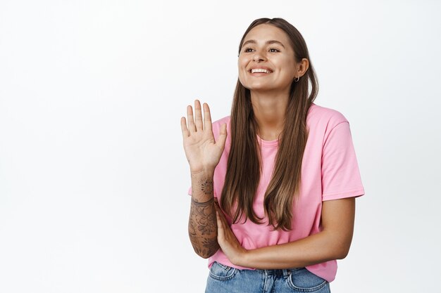 Glückliche junge Frau, die Hand winkt und lächelt, mit freundlichem Gesicht wegschaut, jemanden mit Hallo-Wellen-Geste auf Weiß begrüßt?