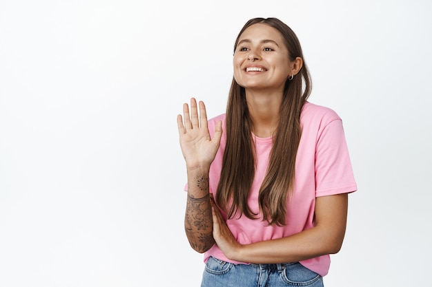 Glückliche junge Frau, die Hand winkt und lächelt, mit freundlichem Gesicht wegschaut, jemanden mit Hallo-Wellen-Geste auf Weiß begrüßt?