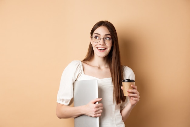 Glückliche junge Frau, die Kaffee trinkt und den Laptop beim Studieren hält und mit fröhlichem Lächeln beiseite schaut ...