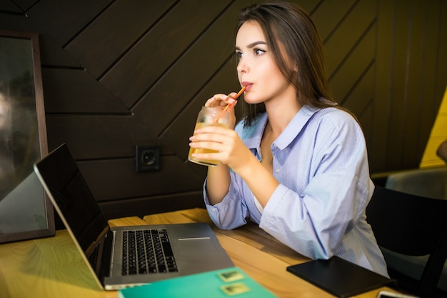 Glückliche junge Frau, die kaltes Getränk im Café mit Laptop trinkt