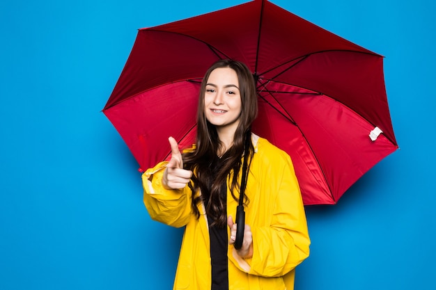 Glückliche junge Frau, die Regenschirm mit gelbem Regenmantel und blauer Wand hält