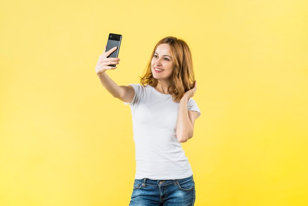Glückliche junge Frau, die selfie am Handy gegen gelben Hintergrund nimmt