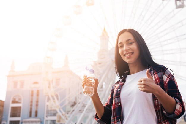 Glückliche junge Frau, die vor Riesenrad steht und Daumen herauf Geste während des Haltens der Wasserflasche zeigt