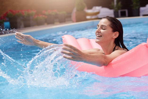 Glückliche junge Frau im Bikini mit aufblasbarer Gummimatratze, spielend und gute Zeit am Wasserbecken während des heißen Sommertages nass