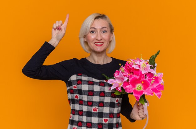 Glückliche junge Frau im schönen Kleid, das Blumenstrauß der Blumen betrachtet, die Front zeigt Zeigefinger lächelnd fröhlich feiern internationalen Frauentag über orange Wand