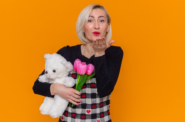 Glückliche junge Frau im schönen Kleid, das Blumenstrauß von Tulpen und Teddybär als Geschenke hält, die Front betrachten, die einen Kuss bläst, der internationalen Frauentag steht über orange Wand steht