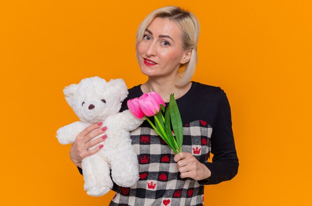 Glückliche junge Frau im schönen Kleid, das Bouquet von Tulpen und Teddybär als Geschenke hält, die Front lächelnd feiern, der internationalen Frauentag steht über orange Wand steht