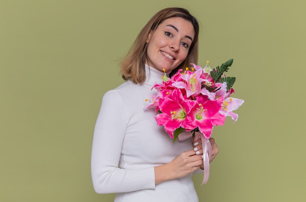 Glückliche junge frau im weißen rollkragenpullover, der blumenstrauß hält, der vorne lächelnd fröhlich feiert internationalen frauentag steht über grüner wand