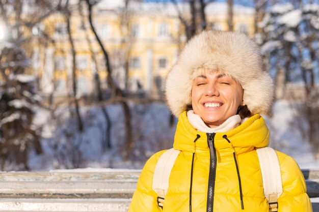 Glückliche junge Frau im Winter in warmer Kleidung in einem schneebedeckten Park an einem sonnigen Tag sitzt auf den Bänken und genießt die frische Luft und den Kaffee allein