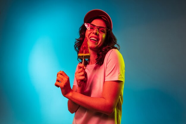 Glückliche junge Frau in der Sonnenbrille, die Süßigkeiten isst und über trendiges blaues Neonstudio lächelt
