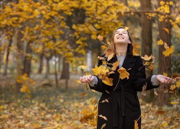 Glückliche junge Frau in einem schwarzen Mantel im Wald unter fliegenden Herbstblättern auf unscharfem Hintergrund, Kopienraum.