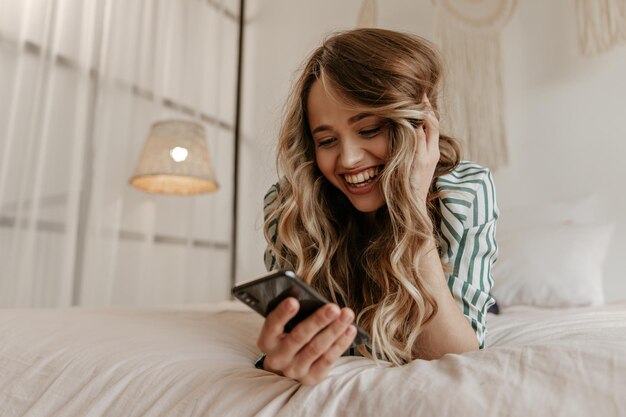Glückliche junge Frau in gestreiftem Seidenhemd liest Nachricht im Telefon und lacht Attraktives Mädchen ruht auf