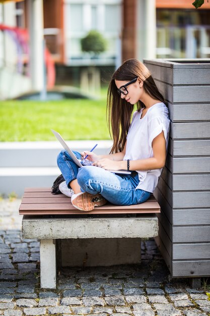 Glückliche junge Frau in lässigem Outfit und Brille mit modernem Laptop und Notizen machen, während auf Bank auf Stadtstraße sitzend