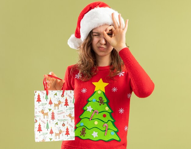 Glückliche junge Frau in rotem Weihnachtspullover und Weihnachtsmütze, die Papiertüte mit Weihnachtsgeschenk hält und zwinkert durch das OK-Zeichen, das über grüner Wand steht?