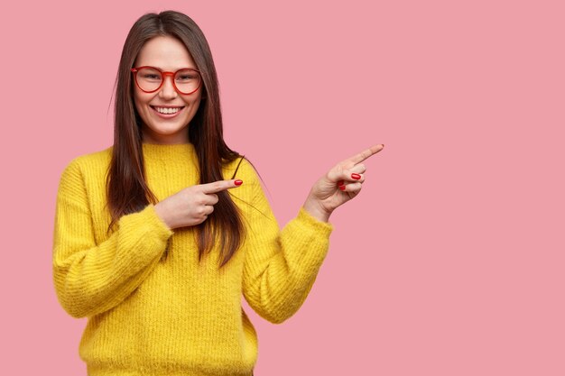 Glückliche junge Frau mit geraden dunklen Haarspitzen beiseite, wirbt für neuen Artikel im Bekleidungsgeschäft, gekleidet in leuchtend gelbem Pullover