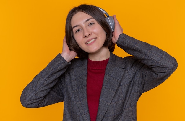 Glückliche junge Frau mit kurzen Haaren, die graue Jacke mit Kopfhörern trägt, die ihre Lieblingsmusik genießt, die fröhlich über orange Wand stehend lächelt