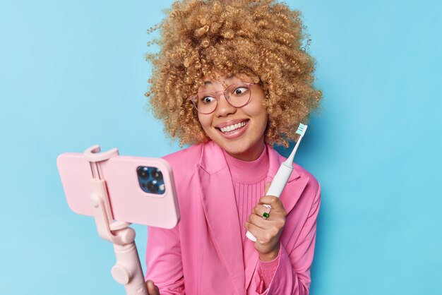 Glückliche junge Frau mit lockigem Haar hält Zahnbürste und Smartphone auf Selfie-Stick und gibt ihren Anhängern Empfehlungen zur Zahnpflege. Sie trägt rosafarbene formelle Kleidung, die über blauem Hintergrund isoliert ist