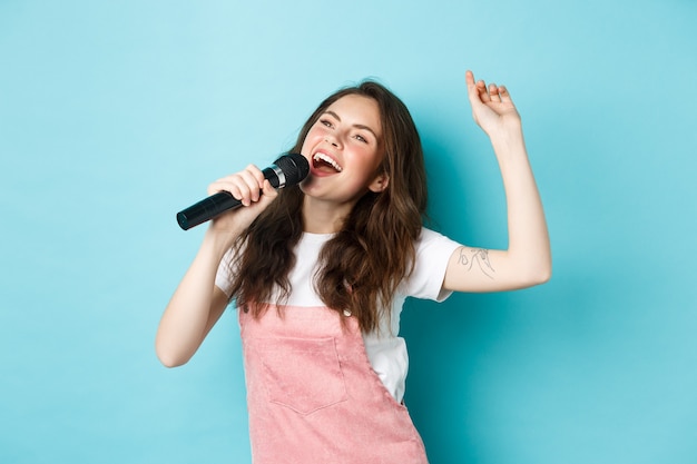 Glückliche junge Frau singt, Sänger hält Mikrofon, tanzt und singt bei Karaoke, stehend über blauem Hintergrund
