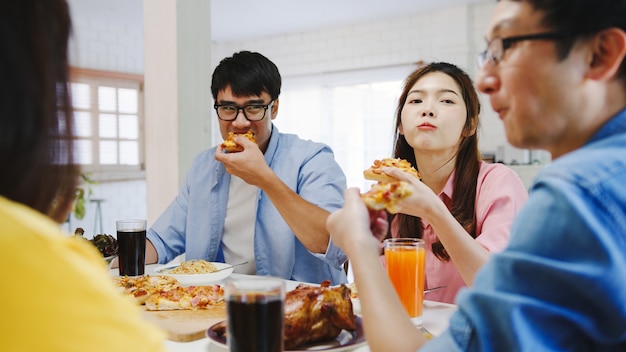 Glückliche junge Freunde Gruppe, die zu Hause zu Mittag isst. Asia Familienfeier, die Pizza Essen isst und lachend das Essen genießt, während sie am Esstisch zusammen im Haus sitzt. Festurlaub und Zusammengehörigkeit.