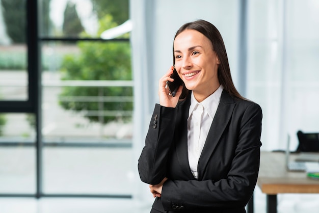 Glückliche junge Geschäftsfrau, die im Büro spricht auf Mobiltelefon steht