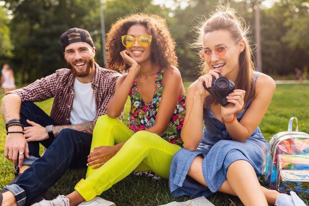 Glückliche junge Gesellschaft der sprechenden lächelnden Freunde, die Park, Mann und Frauen sitzen, die Spaß zusammen haben und mit Kamera reisen