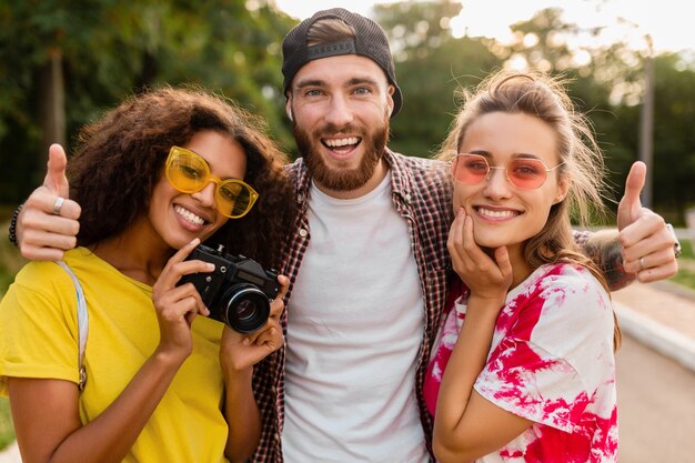 Glückliche junge Gesellschaft von emotional lächelnden Freunden, die im Park mit Fotokamera, Mann und Frauen gehen, die Spaß zusammen haben