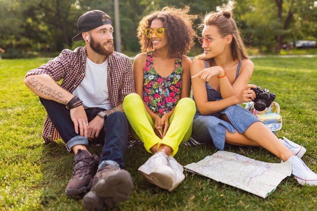 Glückliche junge Gesellschaft von Freunden, die Park, Mann und Frauen sitzen, die Spaß zusammen haben, bunte Sommer-Hipster-Modestil, mit Kamera reisend, sprechend, lächelnd