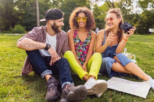 Glückliche junge Gesellschaft von Freunden, die Park, Mann und Frauen sitzen, die Spaß zusammen haben, bunte Sommer-Hipster-Modestil, mit Kamera reisend, sprechend, lächelnd