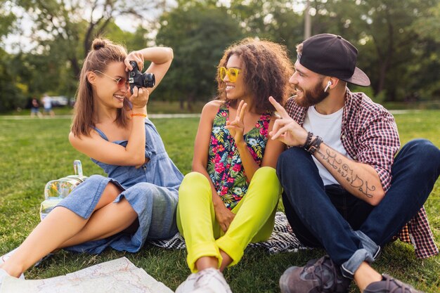 Glückliche junge Gesellschaft von Freunden, die Park, Mann und Frauen sitzen, die Spaß zusammen haben, mit Kamera reisen, Fotos machen