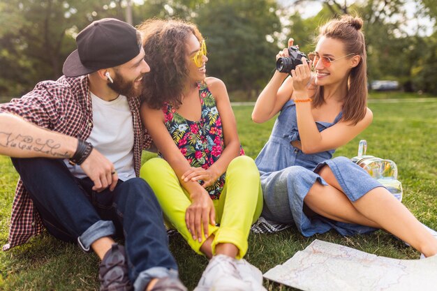 Glückliche junge Gesellschaft von Freunden, die Park sitzen, Mann und Frauen, die Spaß zusammen haben, bunter Sommer-Hipster-Modestil, mit Kamera reisend, offen lachend