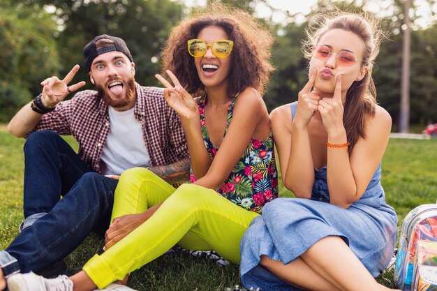 Glückliche junge Gesellschaft von Freunden sitzen Park herumalbern mit verrückten lustigen Gesichtern, Mann und Frauen, die Spaß zusammen haben