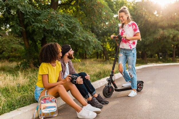 Glückliche junge Gesellschaft von lächelnden Freunden, die im Park auf Gras mit elektrischem Tretroller sitzen, Mann und Frauen, die Spaß zusammen haben